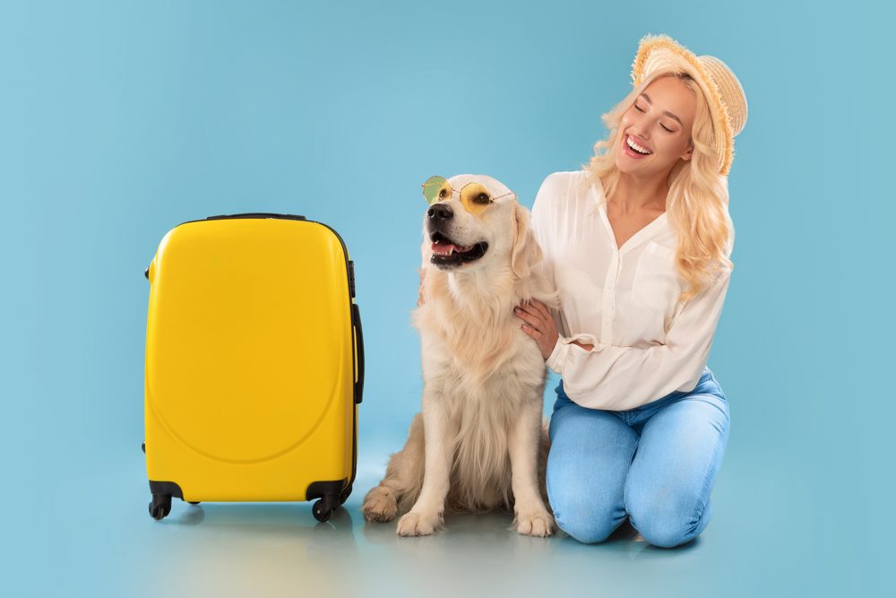A smiling blond woman and golden retriever sit next to bright yellow luggage