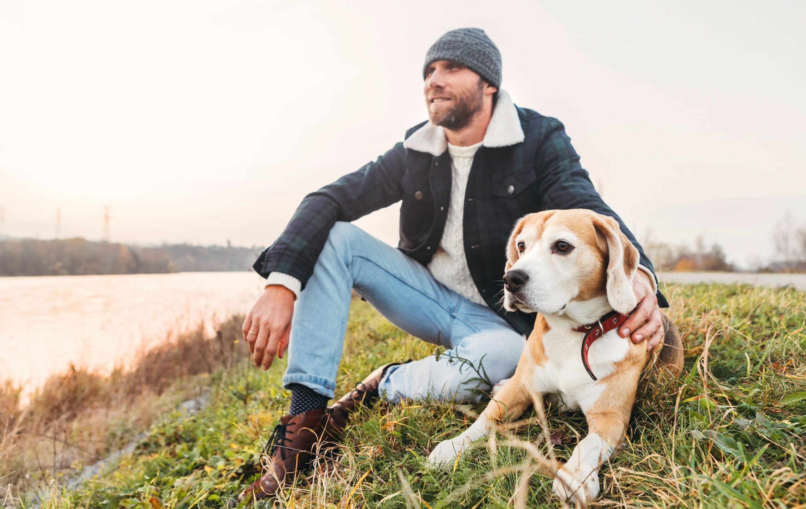 A bearded man in a beanie relaxing on a grassy riverbank next to his emotional support animal in Raleigh, NC.